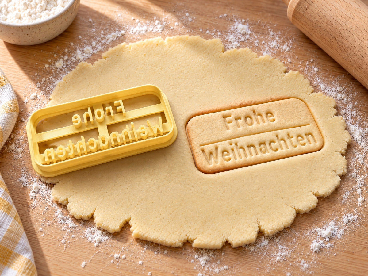 Ausstecher „Frohe Weihnachten“ mit Prägung neben einem rechteckigen Keks mit gerahmtem Weihnachtsgruß auf ausgerolltem Teig