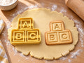 Butter-gelber ABC-Bauklötze-Ausstecher mit Prägung neben Keks mit gestapelten Klötzen A, B und C auf hellem Holz