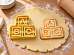 Butter-gelber ABC-Bauklötze-Ausstecher mit Prägung neben Keks mit gestapelten Klötzen A, B und C auf hellem Holz
