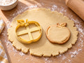 Butter-gelber Apfel-Ausstecher mit Prägung neben gebackenem Keks mit Blatt, Stiel und geschwungenen Rundungslinien auf hellem Holz