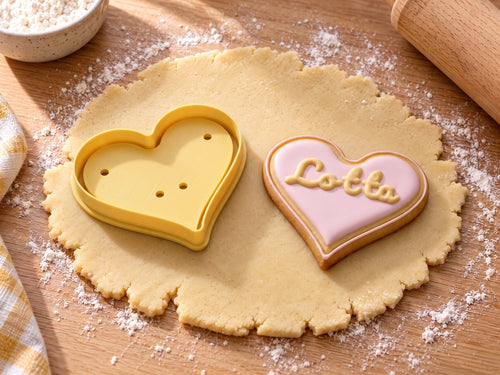 Butter-gelber Herz-Ausstecher neben gebackenem Herzkeks mit rosa Zuckerguss und geprägtem Namen „Lotta“ auf ausgerolltem Teig und hellem Holz