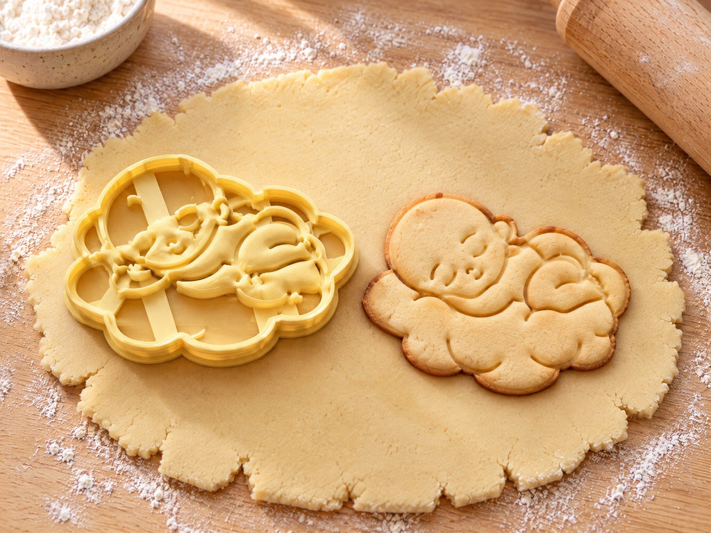 Butter-gelber Ausstecher mit Prägung als Baby auf Wolke neben gebackenem Keks mit schlafendem Baby und bauschiger Wolke auf hellem Holz