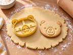 Butter-gelber Bärenkopf-Ausstecher mit Prägung neben gebackenem Keks mit runden Ohren, Punktaugen und Schnäuzchen auf hellem Holz