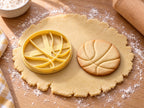 Butter-gelber Basketball-Ausstecher mit Prägung neben rundem gebackenem Keks mit geschwungenen Nahtlinien auf hellem Holz