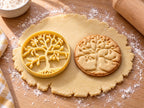 Butter-gelber Lebensbaum-Ausstecher mit Prägung neben rundem Keks mit Krone, Ästen und Wurzeln auf hellem Holz
