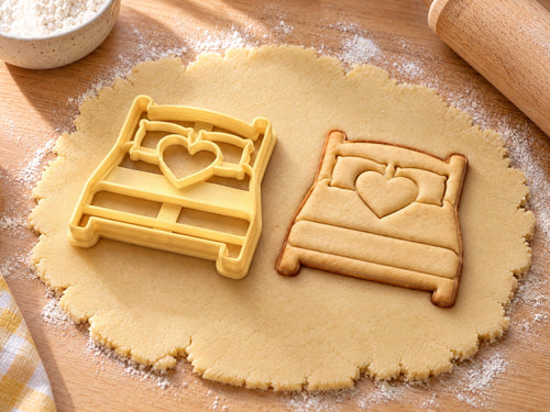 Butter-gelber Bett-Ausstecher mit Prägung neben Keks mit Doppelbett, zwei Kissen und Herz auf hellem Holz
