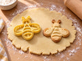 Butter-gelber Bienen-Ausstecher mit Prägung neben gebackenem Keks mit symmetrischer Biene, drei Streifen und zwei Flügeln auf hellem Holz