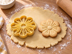 Butter-gelber Blumen-Ausstecher mit Prägung neben gebackenem Keks mit fünf runden Blütenblättern und Staubfäden auf hellem Holz