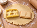 Butter-gelber Buch-Ausstecher mit Prägung neben gebackenem Keks mit aufgeschlagenen Seiten und Lesezeichenband auf hellem Holz