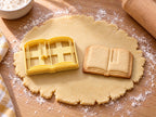 Butter-gelber Buch-Ausstecher mit Prägung neben gebackenem Keks mit aufgeschlagenen Seiten und Lesezeichenband auf hellem Holz