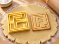 Butter-gelber Ausstecher Buchstabe F mit Prägung neben quadratischem Keks mit F im doppelten Rahmen auf hellem Holz