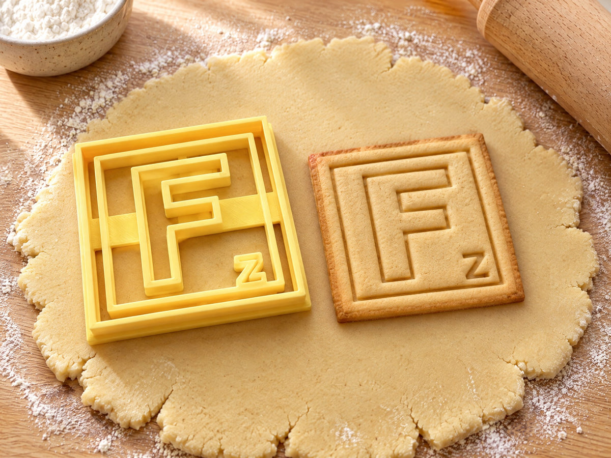 Butter-gelber Ausstecher Buchstabe F mit Prägung neben quadratischem Keks mit F im doppelten Rahmen auf hellem Holz