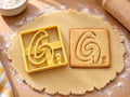 Butter-gelber Ausstecher Buchstabe G mit Prägung neben quadratischem Keks mit spiralförmigem G auf hellem Holz