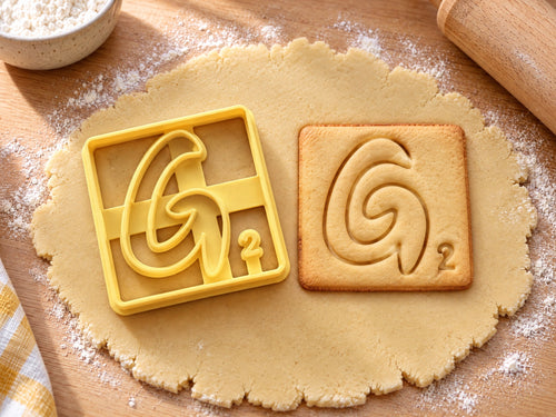 Butter-gelber Ausstecher Buchstabe G mit Prägung neben quadratischem Keks mit spiralförmigem G auf hellem Holz