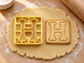 Butter-gelber Ausstecher Buchstabe H mit Prägung neben quadratischem Keks mit verspieltem H und Füßchen auf hellem Holz