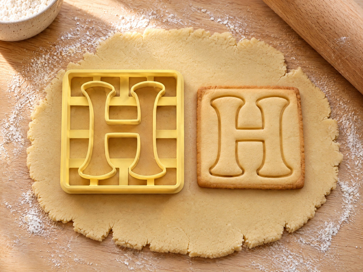 Butter-gelber Ausstecher Buchstabe H mit Prägung neben quadratischem Keks mit verspieltem H und Füßchen auf hellem Holz