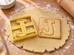 Butter-gelber Ausstecher Buchstabe J mit Prägung neben quadratischem Keks mit geschwungenem J im Plättchen auf hellem Holz