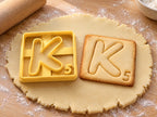 Butter-gelber Ausstecher Buchstabe K mit Prägung neben quadratischem Keks mit gezeichnetem K und Punktwert 5 auf hellem Holz