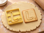 Butter-gelber Ausstecher Buchstabe L mit Prägung neben quadratischem Keks mit L und weichem Bogenfuß auf hellem Holz