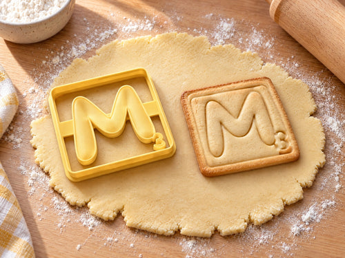 Butter-gelber Ausstecher Buchstabe M mit Prägung neben quadratischem Keks mit weich geschwungenem M auf hellem Holz