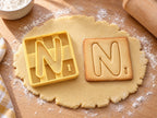 Butter-gelber Ausstecher Buchstabe N mit Prägung neben quadratischem Keks mit rundlichem N und kleiner Zahl auf hellem Holz