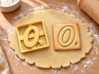 Butter-gelber Ausstecher Buchstabe O mit Prägung neben quadratischem Keks mit kursivem, geneigtem O auf hellem Holz
