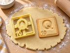 Butter-gelber Ausstecher Buchstabe Q mit Prägung neben quadratischem Keks mit kräftigem Q und Punktwert 10 auf hellem Holz