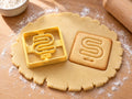 Butter-gelber Ausstecher Buchstabe S mit Prägung neben quadratischem Keks mit geschlängeltem S auf hellem Holz