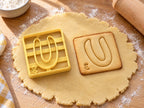 Butter-gelber Ausstecher Buchstabe U mit Prägung neben quadratischem Keks mit rundem U und Punktwert auf hellem Holz