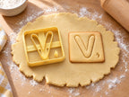 Butter-gelber Ausstecher Buchstabe V mit Prägung neben quadratischem Keks mit weich gerundetem V auf hellem Holz
