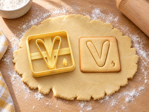 Butter-gelber Ausstecher Buchstabe V mit Prägung neben quadratischem Keks mit weich gerundetem V auf hellem Holz