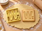 Butter-gelber Ausstecher Buchstabe W mit Prägung neben quadratischem Keks mit breitem, kantigem W auf hellem Holz