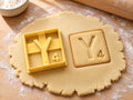Butter-gelber Ausstecher Buchstabe Y mit Prägung neben quadratischem Keks mit Ypsilon und kleiner Vier auf hellem Holz