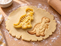 Butter-gelber Dino-Ausstecher mit Prägung neben gebackenem Keks mit grinsendem Saurier, Mini-Ärmchen und Rückenstreifen auf hellem Holz