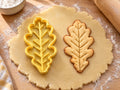 Butter-gelber Eichenblatt-Ausstecher mit Prägung neben gebackenem Keks mit schlanker Form und feinen Seitenadern auf hellem Holz