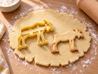 Butter-gelber Elch-Ausstecher mit Prägung neben gebackenem Keks mit Schaufelgeweih und langen Beinen auf hellem Holz
