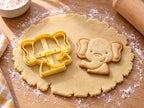 Butter-gelber Elefantenkopf-Ausstecher mit Prägung neben gebackenem Keks mit großen Ohren und erhobenem Rüssel auf hellem Holz