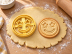 Butter-gelber Emoji-Ausstecher mit Prägung neben rundem Keks mit Sternenaugen, Grinsen und doppeltem Rand auf hellem Holz