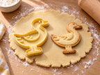 Butter-gelber Enten-Ausstecher mit Prägung neben gebackenem Keks: stehende Ente mit Flügelfedern und Schwimmfüßen auf hellem Holz