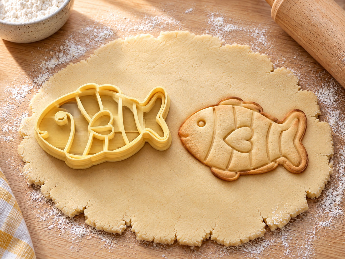 Butter-gelber Fisch-Ausstecher mit Prägung neben gebackenem Keks mit Streifen, Kulleraugen und Herzflosse auf hellem Holz