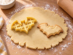 Butter-gelber Fledermaus-Ausstecher neben gebackenem Keks in glatter Silhouette mit Bogenflügeln und Spitzohren auf hellem Holz