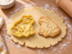 Butter-gelber Frosch-Ausstecher mit Prägung neben gebackenem Keks mit Glubschauge und angewinkeltem Hinterbein auf hellem Holz