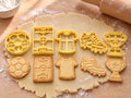 Set Fußball-Fan: 5 butter-gelbe Ausstecher mit Prägung nebeneinander mit gebackenen, geprägten Keksen auf ausgerolltem Teig und hellem Holz