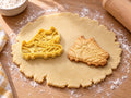 Butter-gelber Ausstecher mit Prägung in Form zweier Fußballschuhe neben gebackenem Keks mit Schnürsenkeln und Stollen auf hellem Holz