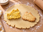 Butter-gelber Ausstecher mit Prägung in Form zweier Fußballschuhe neben gebackenem Keks mit Schnürsenkeln und Stollen auf hellem Holz