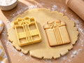 Butter-gelber Geschenk-Ausstecher mit Prägung neben gebackenem Keks: Päckchen von vorn mit Schleife und drei Bändern auf hellem Holz