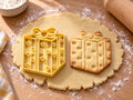 Butter-gelber Geschenk-Ausstecher mit Prägung neben gebackenem Keks: Würfel mit eingeprägten Herzchen und Schleife auf hellem Holz