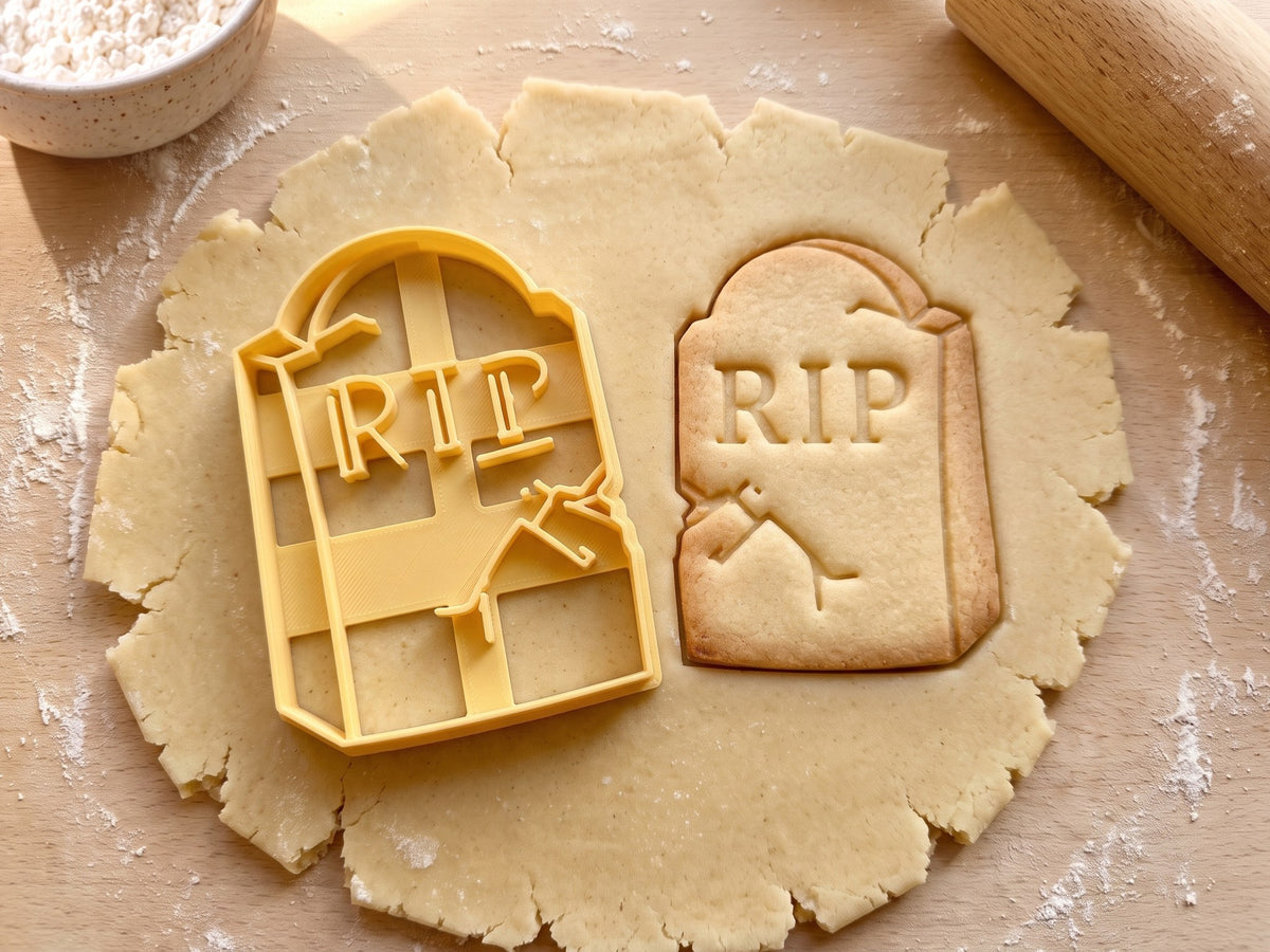 Butter-gelber Grabstein-Ausstecher mit Prägung neben Keks mit RIP-Schriftzug, Riss und abgeplatzter Ecke auf hellem Holz