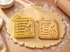 Butter-gelber Haftnotiz-Ausstecher mit Prägung neben gebackenem Keks mit Checkliste, Haken und lachendem Gesicht auf hellem Holz