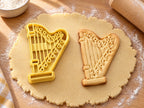 Butter-gelber Harfen-Ausstecher mit Prägung neben gebackenem Keks mit gegliederter Säule, Saiten und Sockel auf hellem Holz